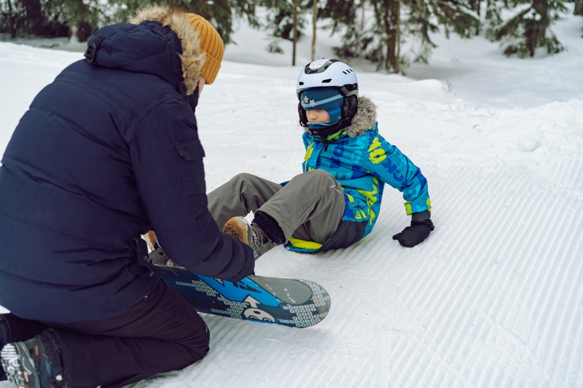 father helping little boy sitting on snow putting his feet in snowboard bindings adjusting straps.